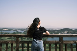 woman in black t-shirt and blue denim jeans standing on brown wooden fence during daytime
