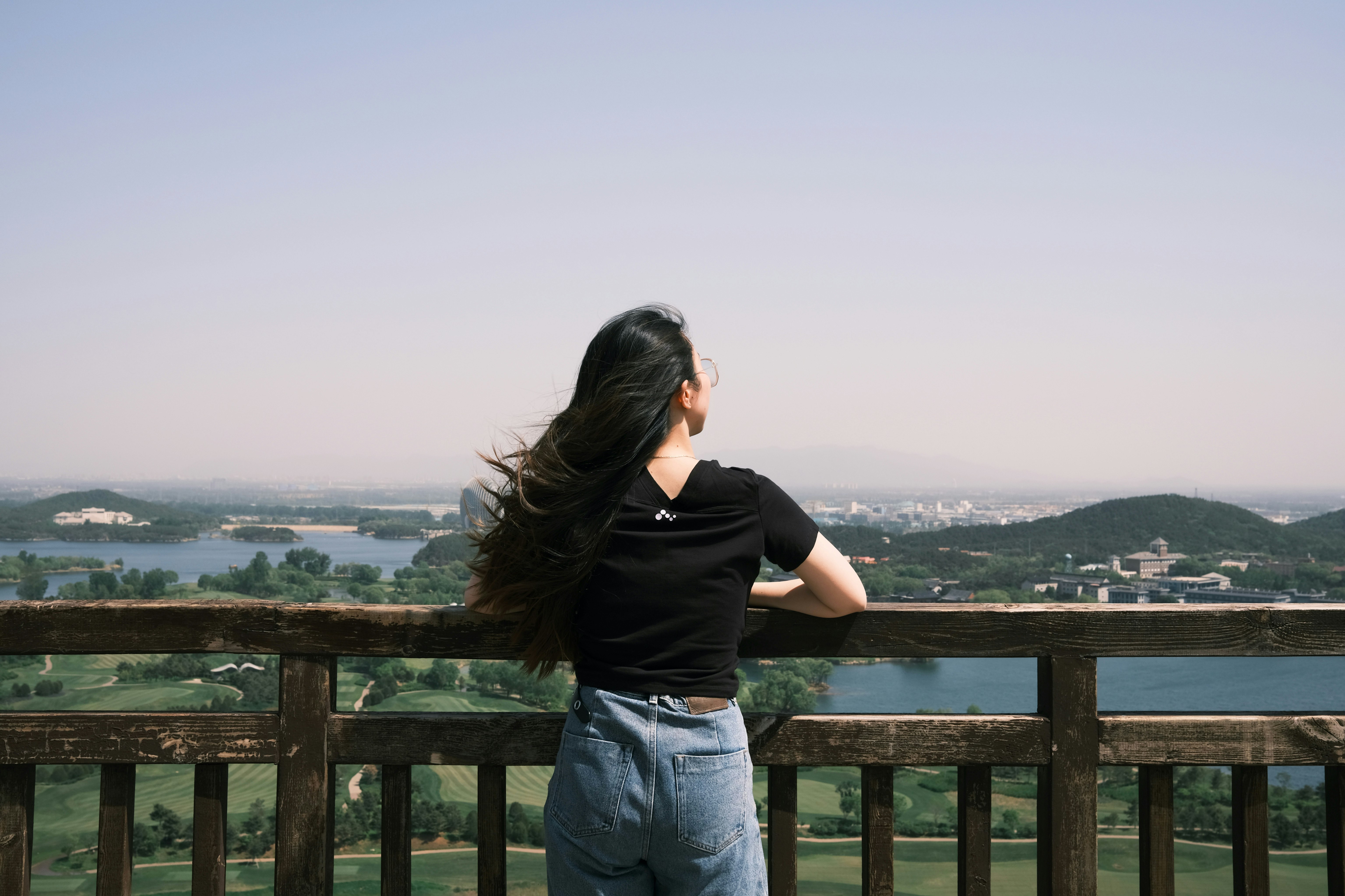 woman in black t-shirt and blue denim jeans standing on brown wooden fence during daytime