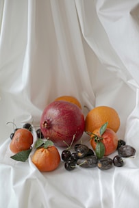A collection of fruits including a large pomegranate, two oranges, small tangerines, dates, and dark cherries is arranged on a white, softly draped fabric background. The fruits are fresh and vibrant, with some small green leaves still attached to the tangerines.