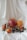 A collection of fruits including a large pomegranate, two oranges, small tangerines, dates, and dark cherries is arranged on a white, softly draped fabric background. The fruits are fresh and vibrant, with some small green leaves still attached to the tangerines.