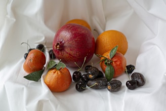 A variety of fruits are arranged on a white draped fabric. A prominent red pomegranate takes center stage, surrounded by oranges and tangerines with green leaves attached. Dark cherries and dried dates are interspersed among the citrus, adding color contrast and texture.