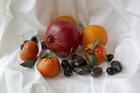 A variety of fruits are arranged on a white draped fabric. A prominent red pomegranate takes center stage, surrounded by oranges and tangerines with green leaves attached. Dark cherries and dried dates are interspersed among the citrus, adding color contrast and texture.