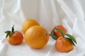 A group of ripe oranges and tangerines rests on a soft, white fabric backdrop. The fruits are vibrant orange, with some adorned with green leaves. The texture of the citrus fruits and their glossy surfaces are highlighted by the soft light.