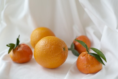 A group of ripe oranges and tangerines rests on a soft, white fabric backdrop. The fruits are vibrant orange, with some adorned with green leaves. The texture of the citrus fruits and their glossy surfaces are highlighted by the soft light.