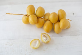 A cluster of yellow dates is resting on a white wooden surface. Two dates are cut open, revealing their inner flesh and seeds.