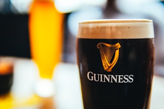 A close-up view of a pint of Guinness with its distinctive dark color and creamy head. The logo and harp emblem are prominently displayed on the glass. In the background, another drink with a lighter, golden hue is slightly blurred, suggesting a depth of focus.