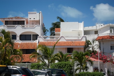 An array of white stucco buildings with red tile roofs surrounded by lush tropical plants and trees. The foreground features multiple parked cars, while the background shows a clear blue sky with some clouds.