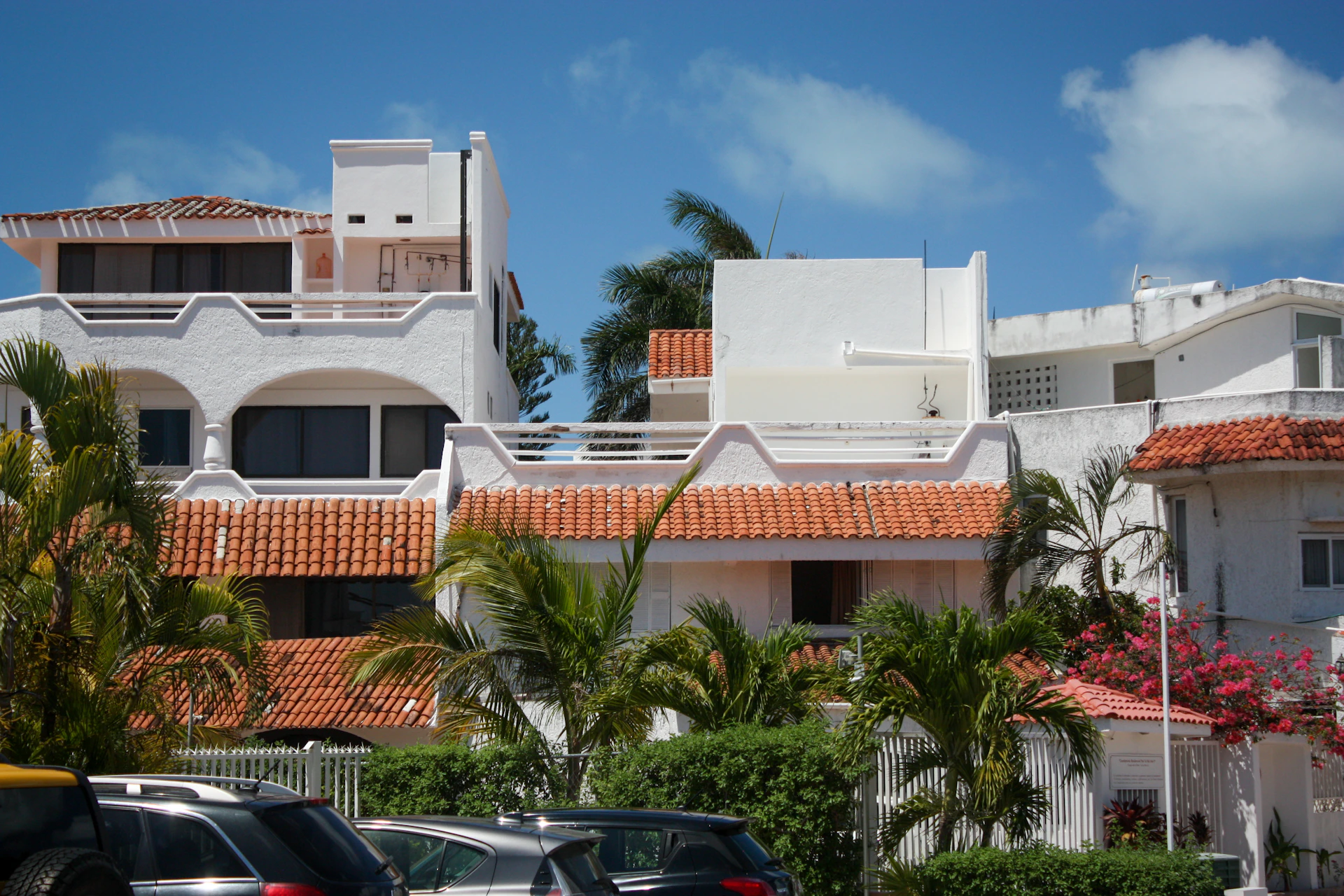 An array of white stucco buildings with red tile roofs surrounded by lush tropical plants and trees. The foreground features multiple parked cars, while the background shows a clear blue sky with some clouds.