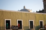 A street scene featuring an olive-green building with four windows, each partially open, and a small balcony lined with plants. Above the building, the top of a historic-looking dome is visible, contrasting with the clear blue sky. A black sign with white letters, 'Yalat Arte Mexicano,' hangs from the balcony, partially obscured by two mannequins wearing colorful clothing.