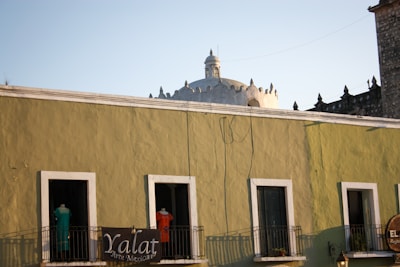 A street scene featuring an olive-green building with four windows, each partially open, and a small balcony lined with plants. Above the building, the top of a historic-looking dome is visible, contrasting with the clear blue sky. A black sign with white letters, 'Yalat Arte Mexicano,' hangs from the balcony, partially obscured by two mannequins wearing colorful clothing.