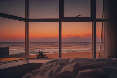 Cozy beachfront hotel room with large windows showing ocean view at sunset.