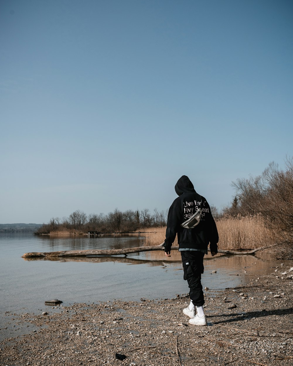 homme en sweat à capuche noir debout sur un champ brun près du lac pendant la journée