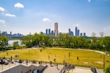 A park with a large grassy area where people are walking, sitting, and playing. Trees surround the park and there is a river visible in the background. The city's skyline with modern high-rise buildings is also prominently visible, with a bright blue sky overhead and a few scattered clouds.