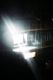 A car headlight shines brightly in the darkness, casting a wide beam of light. Raindrops are visible, illuminated by the light, creating streaks across the image. The scene conveys a sense of motion and focus amidst a dark, rainy night.