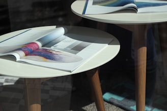 An arrangement of two small round tables made of wood and topped with glossy magazines, open to pages featuring colorful abstract art and design elements. Natural light casts soft shadows, emphasizing the textures of the tables and the sheen of the magazine pages.