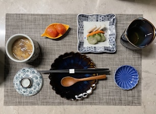 A neatly arranged Japanese-style meal presented on a woven placemat. The setting includes a cup of soup with a patterned lid, a plate with assorted sushi, a cup of tea, sliced pickled vegetables in a small dish, and chopsticks resting on a blue, flower-shaped plate alongside a wooden spoon.