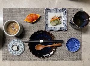 A neatly arranged Japanese-style meal presented on a woven placemat. The setting includes a cup of soup with a patterned lid, a plate with assorted sushi, a cup of tea, sliced pickled vegetables in a small dish, and chopsticks resting on a blue, flower-shaped plate alongside a wooden spoon.