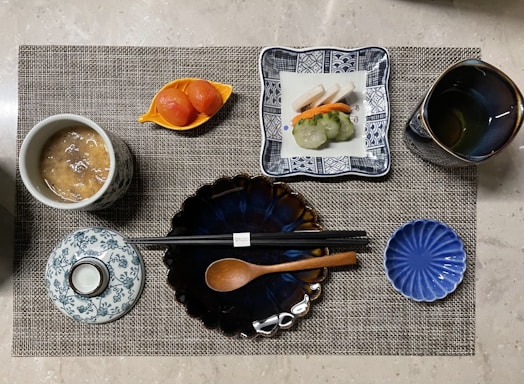 A neatly arranged Japanese-style meal presented on a woven placemat. The setting includes a cup of soup with a patterned lid, a plate with assorted sushi, a cup of tea, sliced pickled vegetables in a small dish, and chopsticks resting on a blue, flower-shaped plate alongside a wooden spoon.