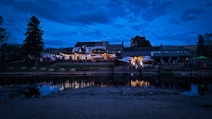 A cozy riverside building illuminated by warm lights during the evening, with tables and umbrellas set up on an outdoor patio. The calm water reflects the vibrant lights and the dark silhouettes of trees and hills create a serene backdrop.