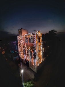 A house is adorned with festive lights, including strands of orange and white bulbs, beautifully illuminating the exterior against the backdrop of a dark sky. The lights highlight the architectural details of the building, including its windows and roof, creating a warm and inviting atmosphere. Shadows and soft lighting enhance the ambiance.
