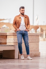 A man stands casually against a stone bench with intricate patterns, wearing a tan jacket over a white T-shirt, blue jeans, and white sneakers. The background features a blurred outdoor space with neutral tones and architectural elements.