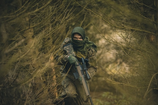 A person dressed in camouflage attire is navigating through a dense forest. The individual is holding a rifle and wearing a balaclava that covers most of their face, blending into the surrounding wilderness. The atmosphere is tense and focused, with branches and leaves obscuring parts of the view.