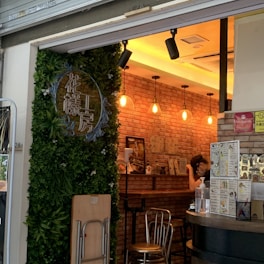 An inviting cafe entrance with a lush green wall and signage featuring Chinese characters. The interior showcases a warm and cozy atmosphere with brick walls, hanging lights, and a wooden counter. A person is seen inside, possibly a staff member at work. Menus and other promotional materials are displayed on the walls.