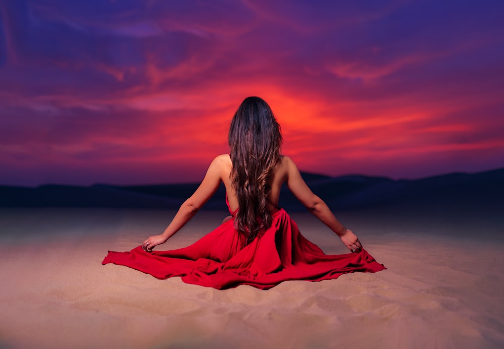 woman in red dress sitting on sand during sunset