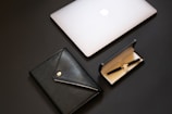 A sleek and modern composition featuring a closed silver laptop with an apple logo, a black leather portfolio with a button closure, and a black fountain pen inside an open black case with a beige interior.