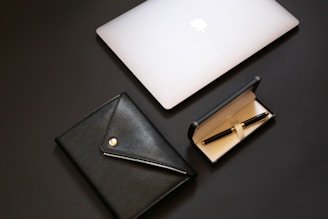 A sleek and modern composition featuring a closed silver laptop with an apple logo, a black leather portfolio with a button closure, and a black fountain pen inside an open black case with a beige interior.