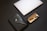 A sleek and modern composition featuring a closed silver laptop with an apple logo, a black leather portfolio with a button closure, and a black fountain pen inside an open black case with a beige interior.