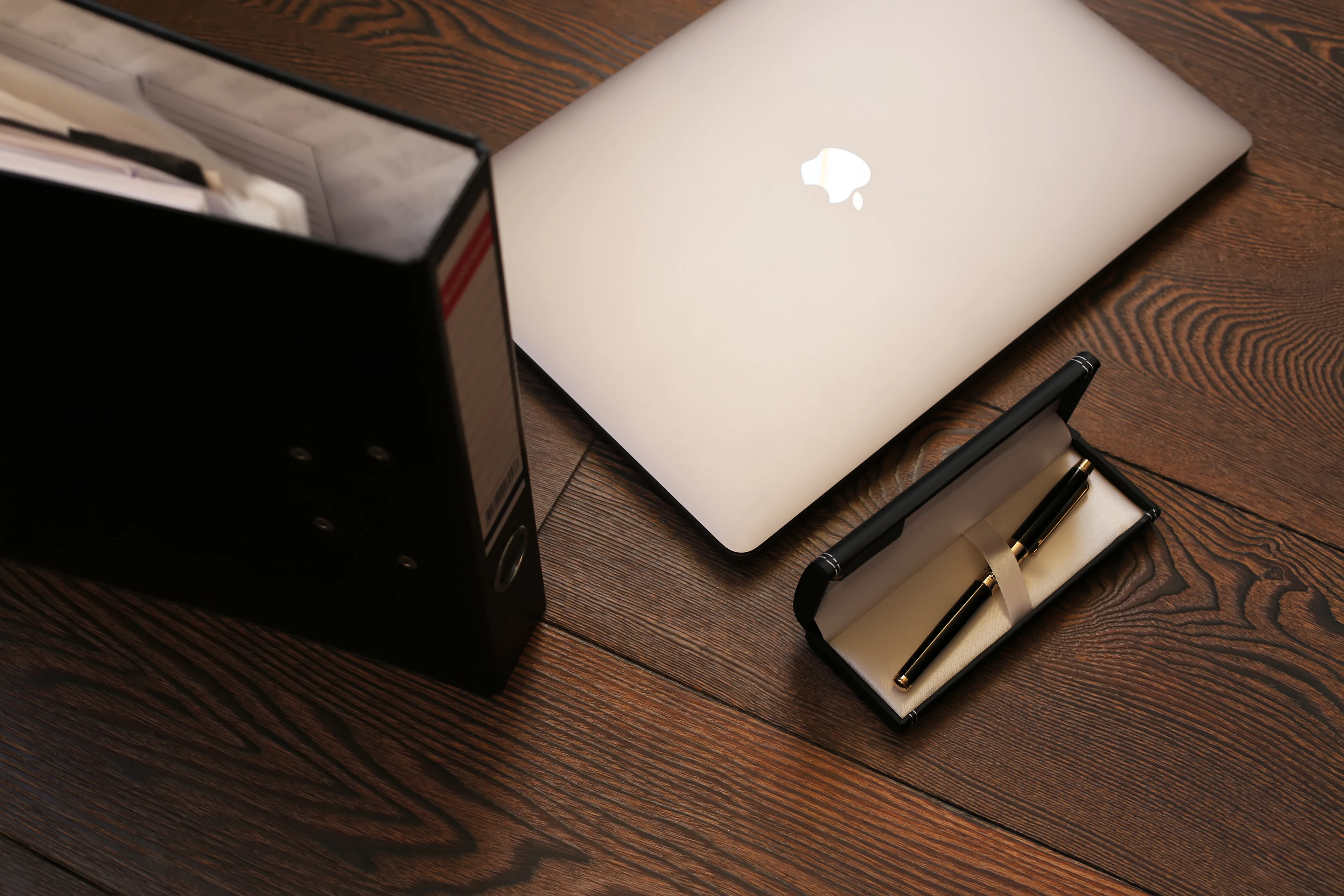 A closed silver laptop on a dark wooden floor beside a black ring binder and an open pen case
