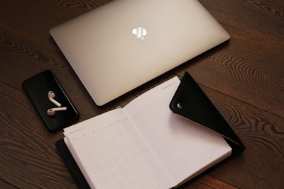 A laptop with a glowing logo is placed on a wooden surface next to a smartphone with wireless earbuds. An open planner with a zippered cover lies beside the devices.