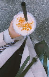 A hand is holding a cup of bubble tea with a dark straw and orange boba pearls on top. The person is dressed in a light-colored outfit with a plaid pattern visible on the inner layer. A green bag with a strap is also in view, and the background appears to be a textured surface, possibly pavement.