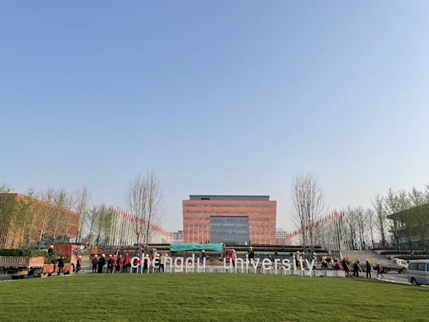 A large red brick building is seen in the center, flanked by rows of trees and a neatly manicured lawn in the foreground. Several workers in uniform are gathered near vehicles and are possibly engaging in landscaping activities. The letters 'Chengdu University' are prominently displayed on the lawn in front.