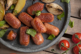 A plate containing cooked sausages garnished with green herbs, alongside roasted onions and potato wedges. Cherry tomatoes are placed nearby on a rustic wooden surface.