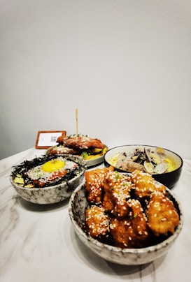 A collection of appetizing dishes displayed on a marble table, featuring a bowl of glazed fried chicken pieces sprinkled with sesame seeds, a bowl of rice topped with seaweed, an egg yolk, and sauce, a dish containing shellfish over pasta, and a plate of seasoned meat on lettuce.