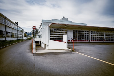 A fire station with the name Feuerwehr Risch is pictured, featuring multiple garage doors lined up along the building. The scene is set on a cloudy day with overcast skies. There is a road leading up to the building, with a no-entry sign visible. To the left, an older industrial building is partially visible, showing signs of wear. The ground is paved, and there are sparse patches of grass.
