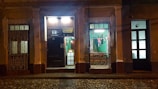 An illuminated vintage facade of a restaurant with ornate ironwork, displaying a Cuban flag in the window. The street is dimly lit, highlighting the rustic charm of the building. There are signs indicating the restaurant's name and address above a dark wooden door, with a warmly lit interior visible through the window.