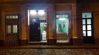 An illuminated vintage facade of a restaurant with ornate ironwork, displaying a Cuban flag in the window. The street is dimly lit, highlighting the rustic charm of the building. There are signs indicating the restaurant's name and address above a dark wooden door, with a warmly lit interior visible through the window.