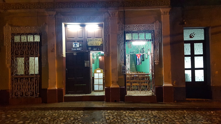An illuminated vintage facade of a restaurant with ornate ironwork, displaying a Cuban flag in the window. The street is dimly lit, highlighting the rustic charm of the building. There are signs indicating the restaurant's name and address above a dark wooden door, with a warmly lit interior visible through the window.