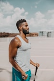 A man is standing outdoors in a spacious area with a clear blue sky. He is wearing a light grey sleeveless shirt and turquoise shorts, and is holding a skipping rope. The background includes a blurred view of a building and some trees.