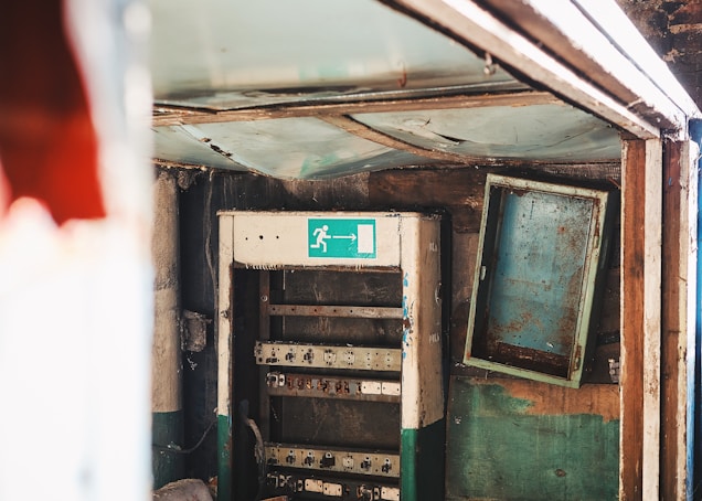 An old, rusted electrical or communication panel is situated in a worn-out structure. The panel is open and mounted on a wall with peeling paint. Above the panel, a faded emergency exit sign with an arrow and figure indicates an escape route. The surrounding area shows signs of neglect and decay, with a dusty, cluttered environment.