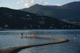 A calm lake with a curving sandbar extending into the water, where several people and a dog are wading and walking. The background features lush, green mountains under a sky dotted with a few clouds, and hillsides scattered with houses.
