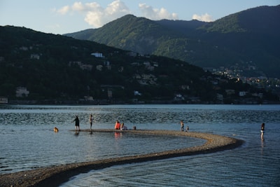 A calm lake with a curving sandbar extending into the water, where several people and a dog are wading and walking. The background features lush, green mountains under a sky dotted with a few clouds, and hillsides scattered with houses.