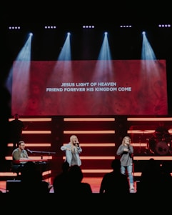 A stage with musicians performing, highlighted by bright lights and a large screen displaying lyrics: 'Jesus light of heaven, friend forever his kingdom come'. Two singers are standing in the center, while a musician plays a keyboard on the left. A drummer is positioned on the right behind a drum set.
