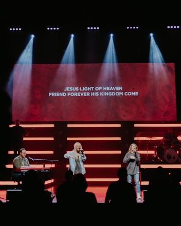 A stage with musicians performing, highlighted by bright lights and a large screen displaying lyrics: 'Jesus light of heaven, friend forever his kingdom come'. Two singers are standing in the center, while a musician plays a keyboard on the left. A drummer is positioned on the right behind a drum set.