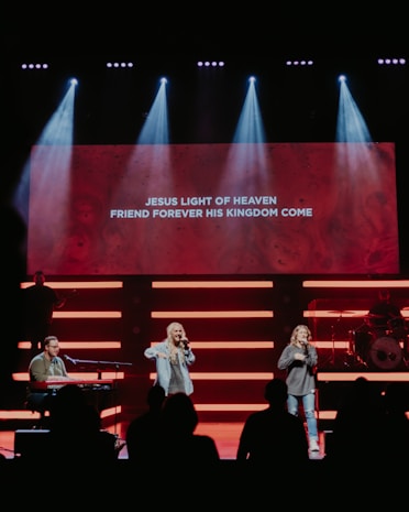 A stage with musicians performing, highlighted by bright lights and a large screen displaying lyrics: 'Jesus light of heaven, friend forever his kingdom come'. Two singers are standing in the center, while a musician plays a keyboard on the left. A drummer is positioned on the right behind a drum set.