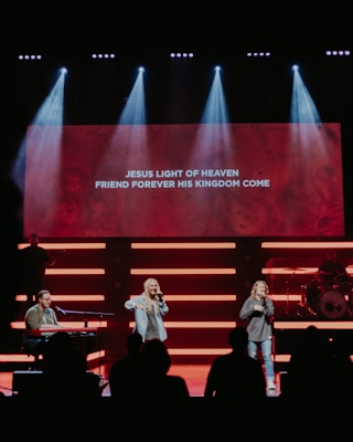 A stage with musicians performing, highlighted by bright lights and a large screen displaying lyrics: 'Jesus light of heaven, friend forever his kingdom come'. Two singers are standing in the center, while a musician plays a keyboard on the left. A drummer is positioned on the right behind a drum set.