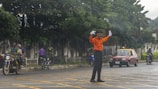 A traffic officer wearing an orange uniform stands in the middle of a road with arms raised, likely directing traffic. Nearby, a red car and several motorcyclists are visible. The background is lined with lush green trees and a fence.